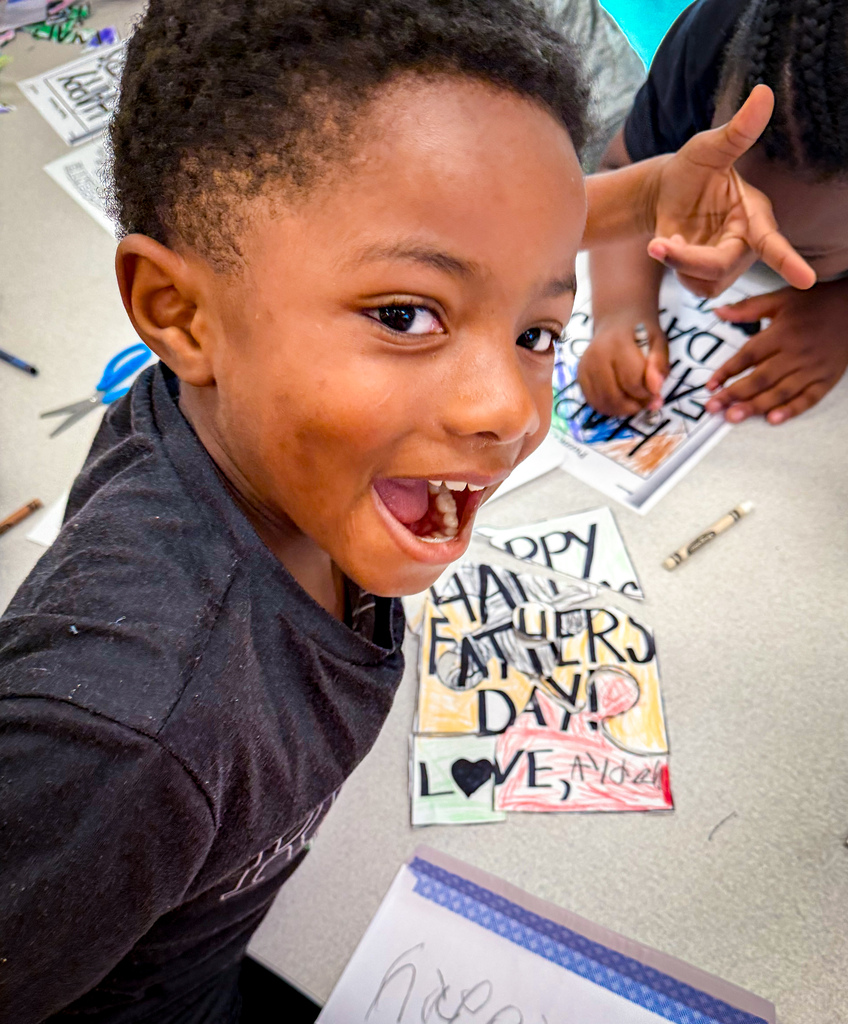 A smiling young boy with a dark t-shirt. Behind him, there are two children, one holding scissors, crafting cards.