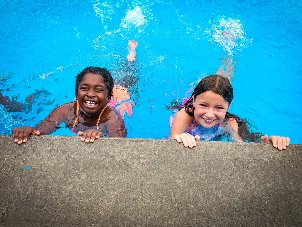 Three children swimming in a pool. Two lean on a concrete edge; one smiles. Background is blue.
