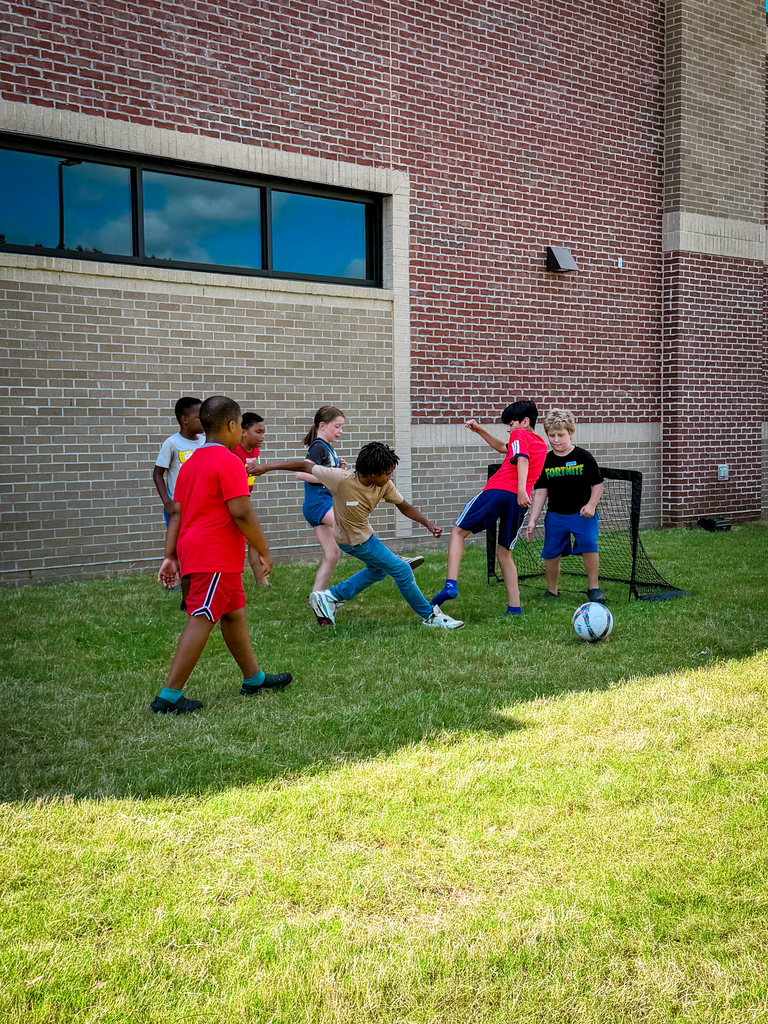 A group of children playing soccer in front of a brick building. Some wear shorts and t-shirts.