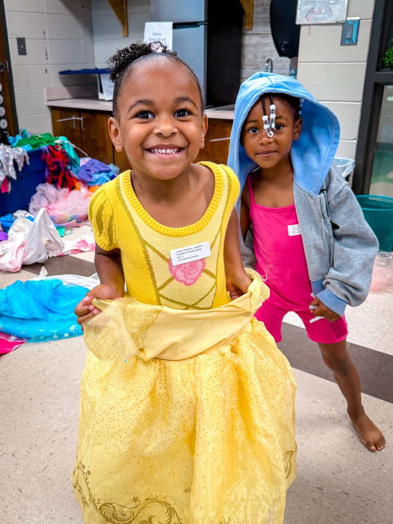 Two children smile, one in a yellow dress and the other in pink shorts and a gray hoodie.