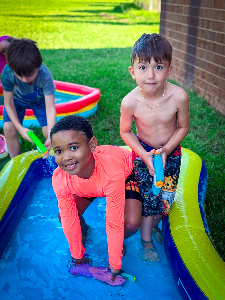 Three young boys play in a small pool. One kneels and holds a toy. Two others stand behind.