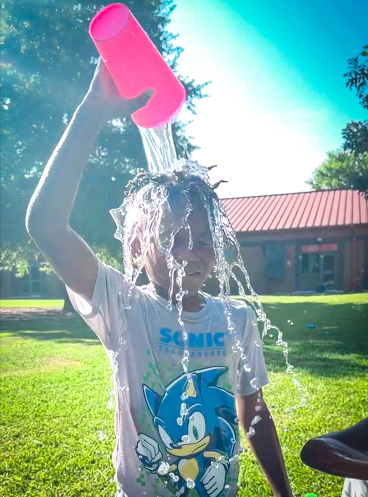 A child pours water from a pink cup over their head on a sunny day in a park.