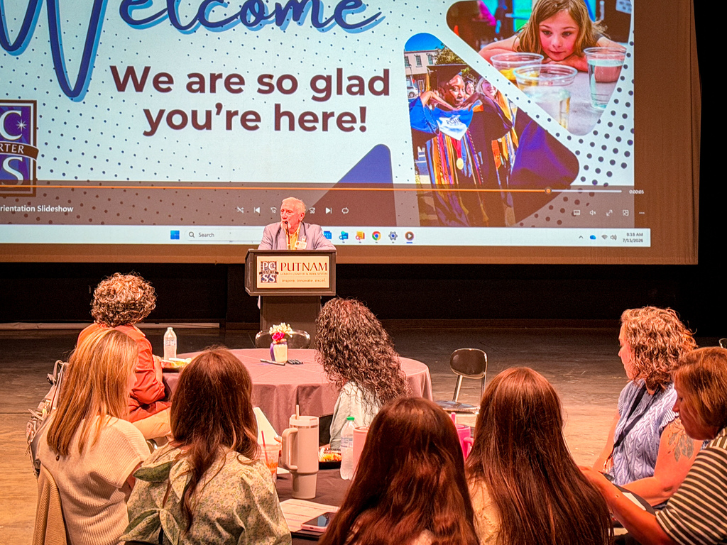 People sit at tables, facing a stage. A speaker stands at a podium. A projector screen displays a welcoming message.
