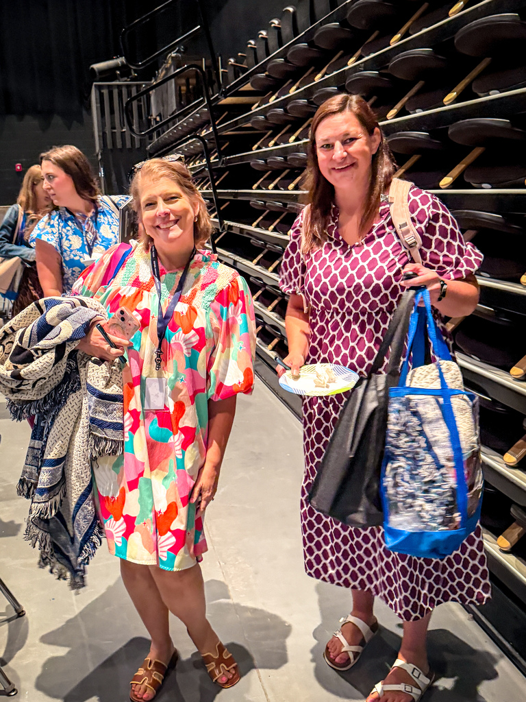 Two women stand in a room with a wall of shoes, one holding a bag and plate, and the other holding a bag.