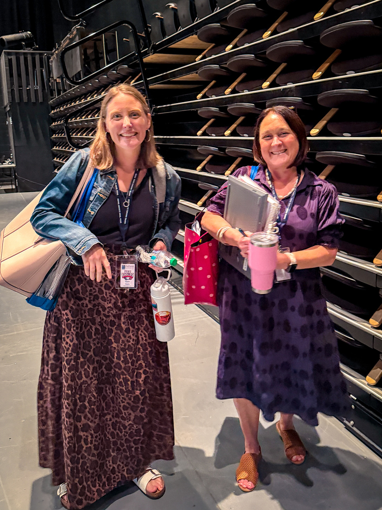 Two women stand in a theater aisle; one wears a leopard skirt, the other a purple dress. Both carry bags and cups.