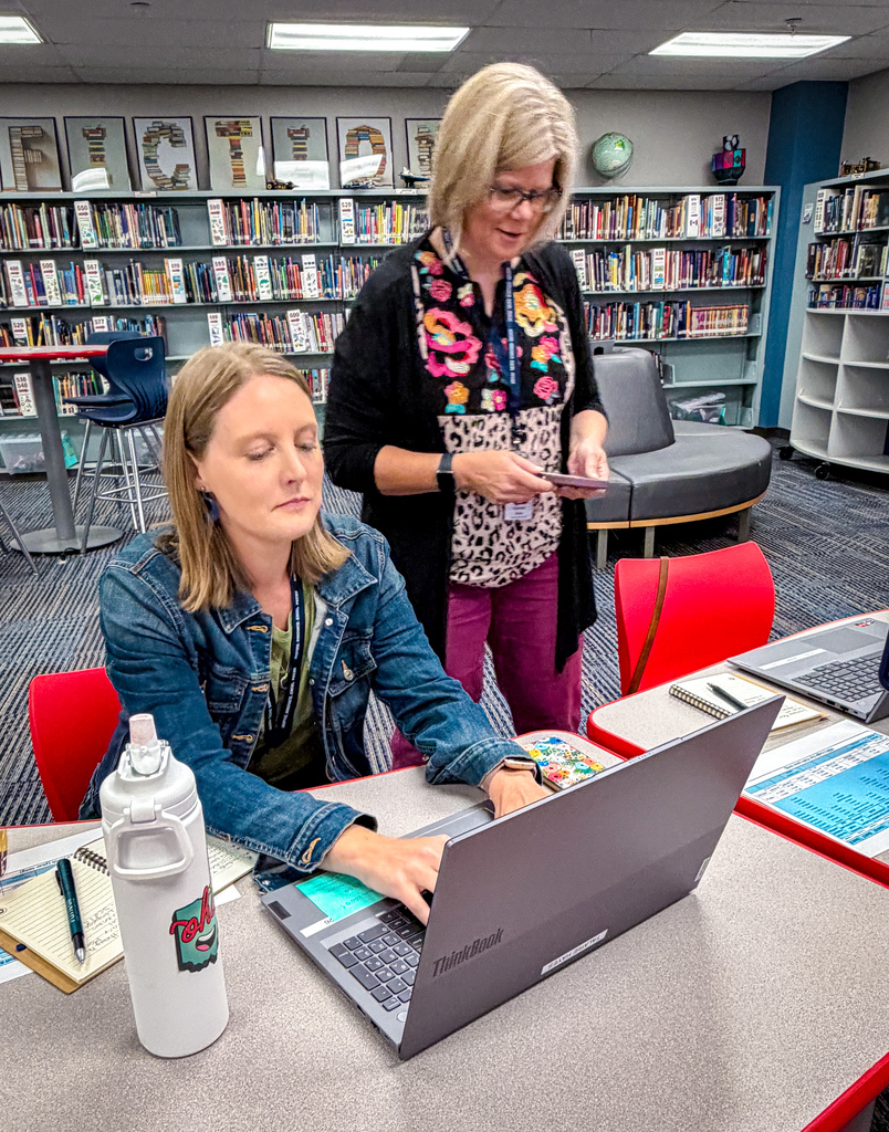 Two women work on a laptop at a library table. One woman wears glasses and a watch.