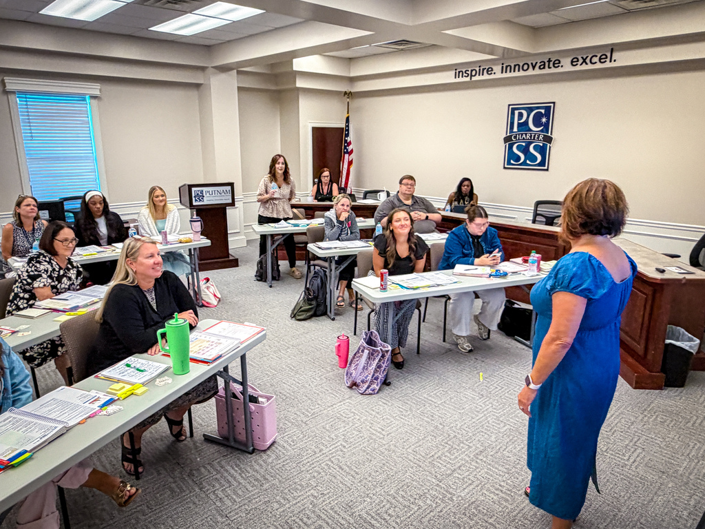 A classroom with students seated at tables, one woman in blue stands at the front. A banner says "Imagine Innovate Excel".