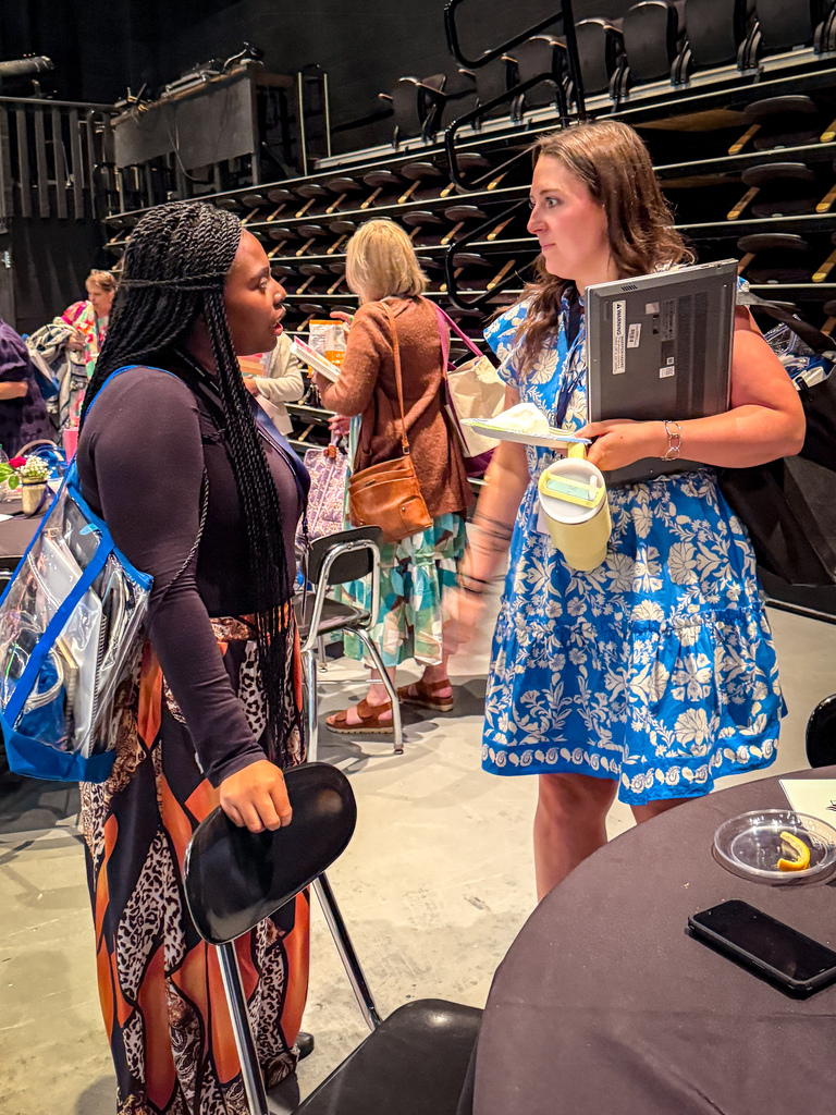 Two women converse in a room with tables, chairs, and rows of seats in the background.