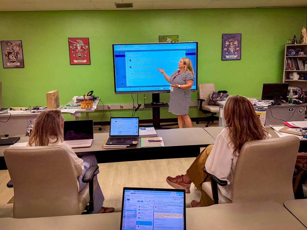 A woman stands in front of a large screen, teaching in a room with a green wall. Two women sit at a table with laptops.