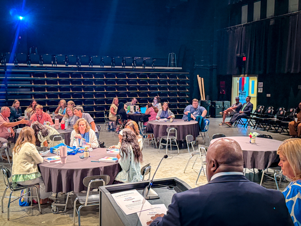 People seated around tables in a large room, with a man at a podium facing the attendees.