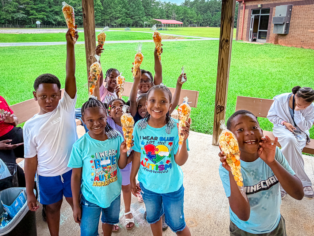 Children holding food, smiling and standing outside, one girl with a rainbow shirt, green grass, and building.