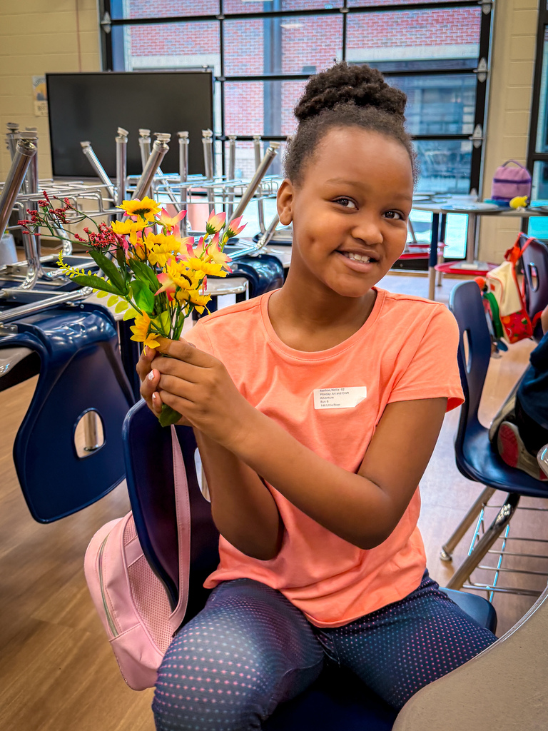 A young girl with a smile holds a bouquet of yellow flowers, sitting in a classroom.