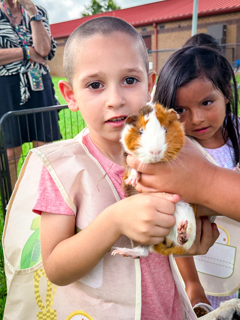 Two children holding a guinea pig, one wearing a pink shirt. Behind them, a person stands.