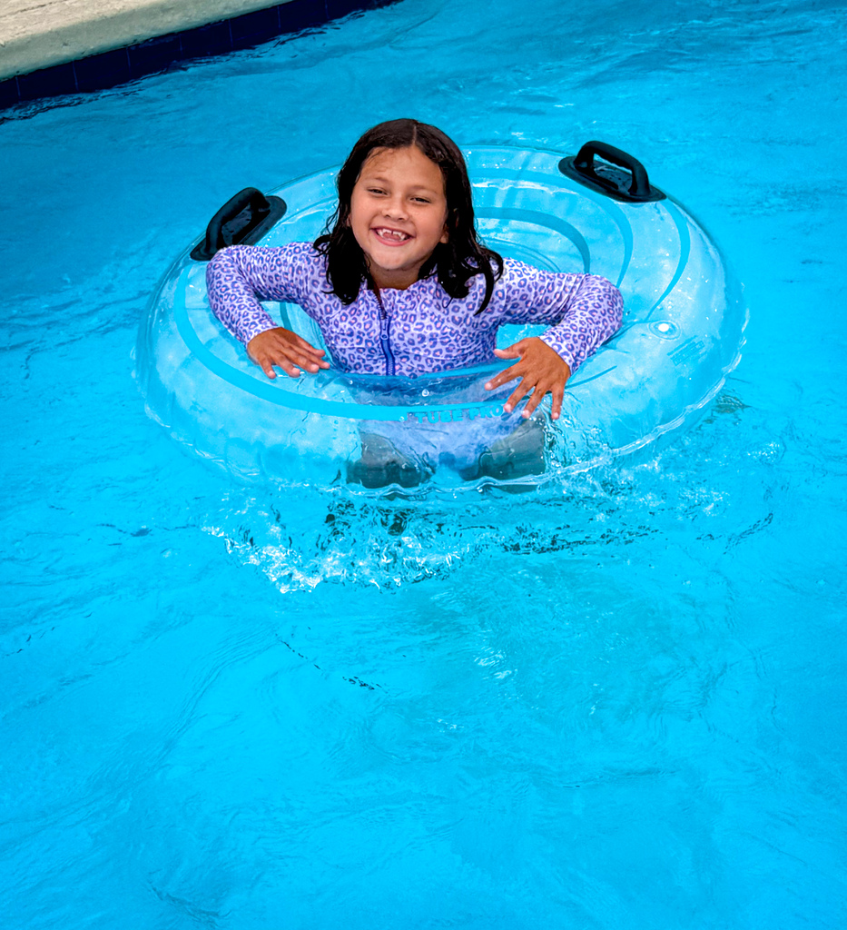 A young girl in a blue float tube, smiling, floats in a swimming pool.