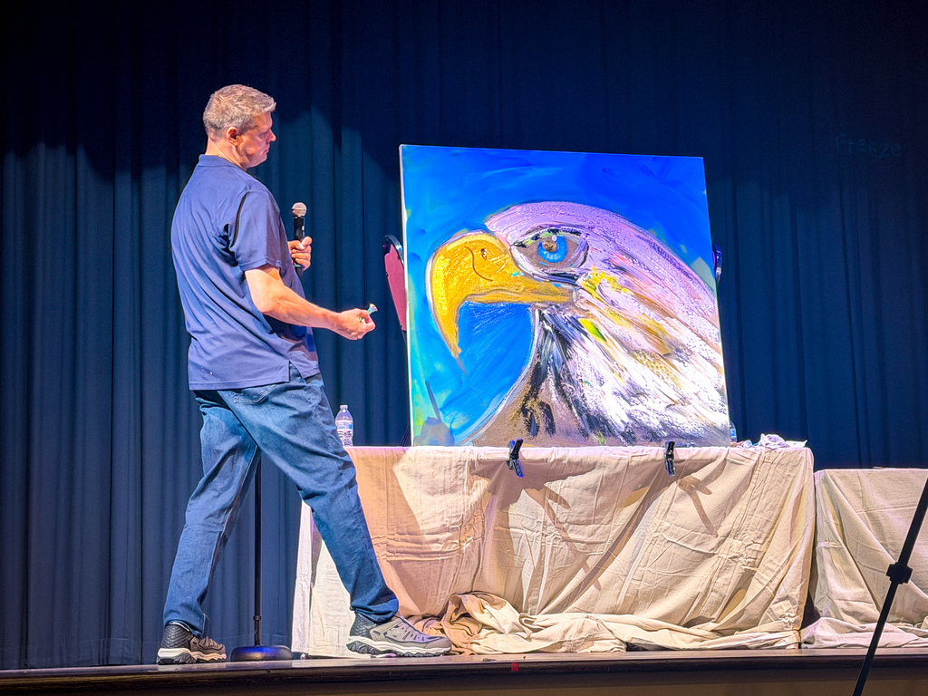 Person on stage with a microphone, painting a bald eagle on a large canvas. The stage has a blue curtain backdrop.