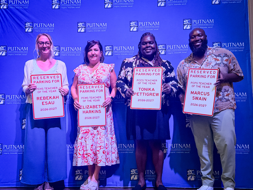 Four individuals stand in front of a blue backdrop, each holding a white sign with black text. The signs read "Reserved Parking" and names.