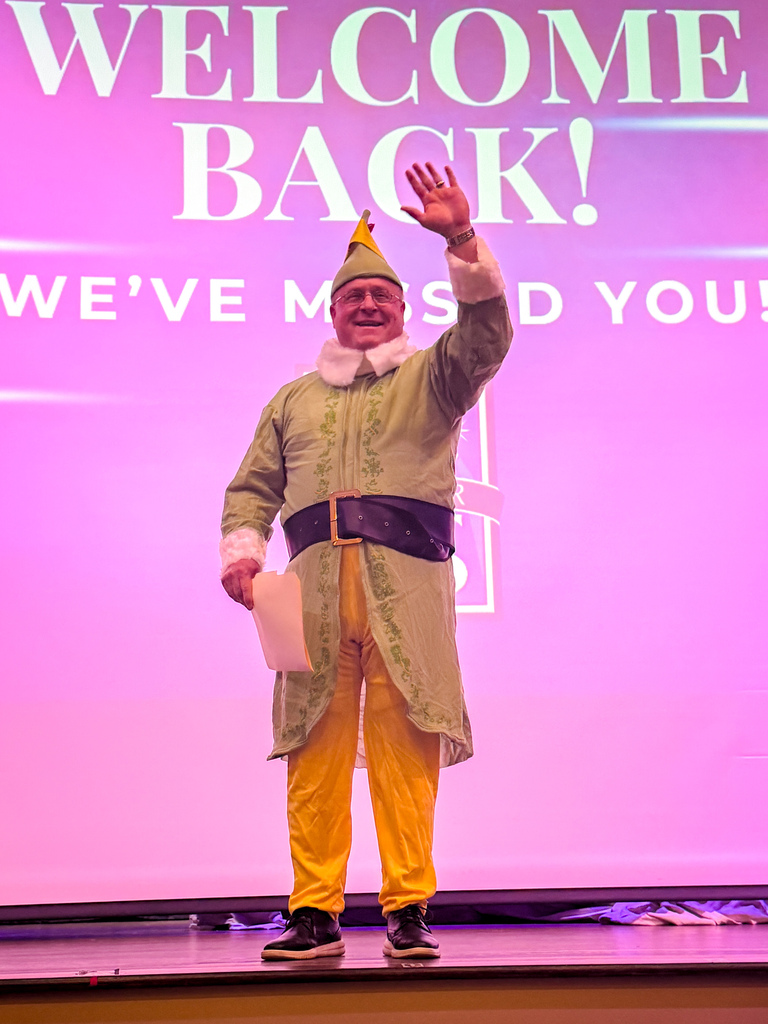 A person dressed as an elf waving at an audience with a pink screen behind them that reads "Welcome Back! We've missed you!"
