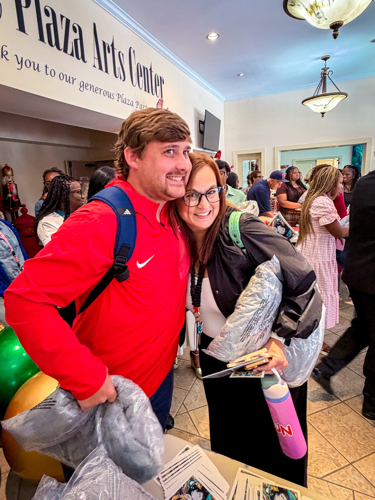 A man and woman stand inside, smiling, with the man holding bags and a backpack. Behind them, people walk in a hallway with a tiled floor.