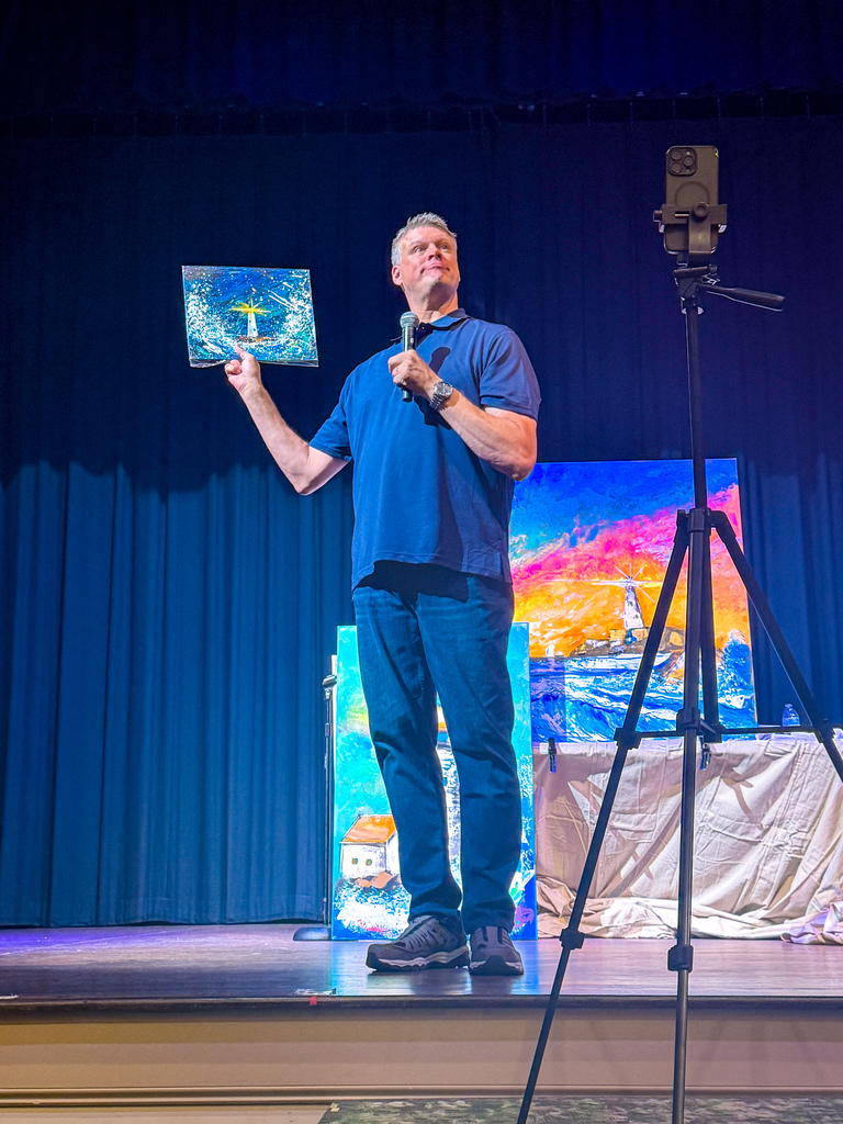 Person on stage holding a framed picture, speaking into a microphone. Blue curtains, a painting, and a tripod behind.