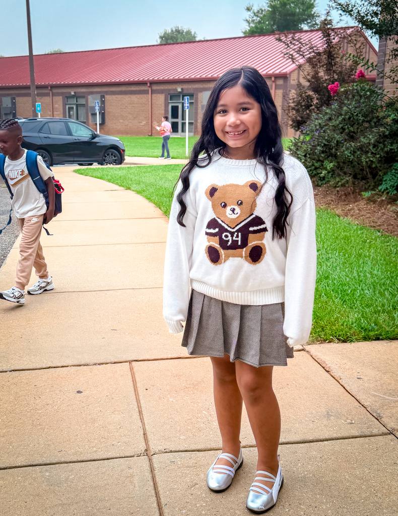 A girl with a bear sweater stands on a sidewalk outside a building. Behind her, a person walks with a backpack.