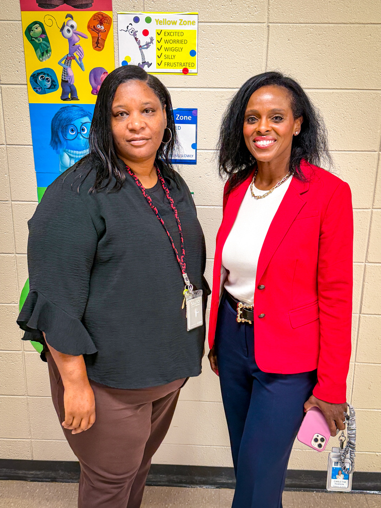 Two women stand in front of a wall with educational posters, smiling. One wears a red blazer, the other a black blouse.
