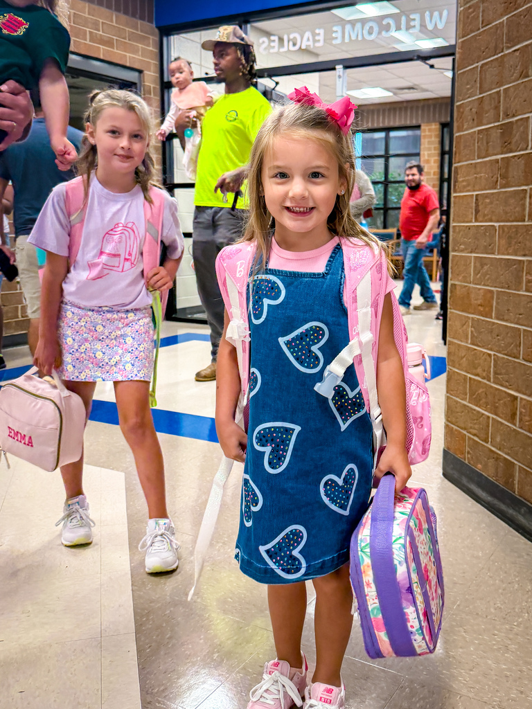 Two young girls in a hallway, one with a heart-patterned dress and the other with a pink backpack.