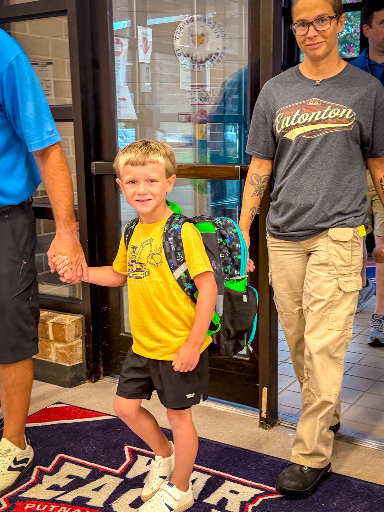 A child in a yellow t-shirt holds hands with a man in a gray shirt. Both wear backpacks. A doorway and rug with text are in the background.