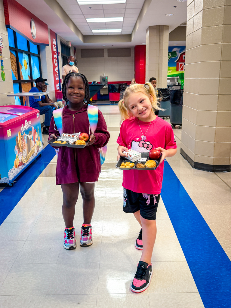 Two girls in a school hallway carrying trays of food. One wears a Hello Kitty shirt, the other a maroon dress.
