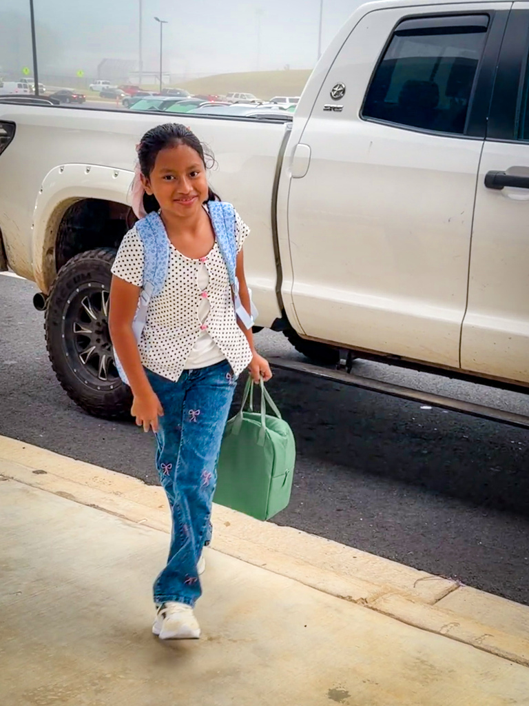 Young girl with backpack holds a green bag, walking on sidewalk with white truck in background.