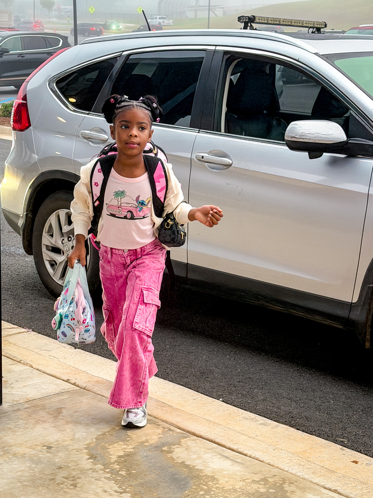 A child wearing a pink shirt, pink pants, and a backpack, walks on the sidewalk near a silver car.