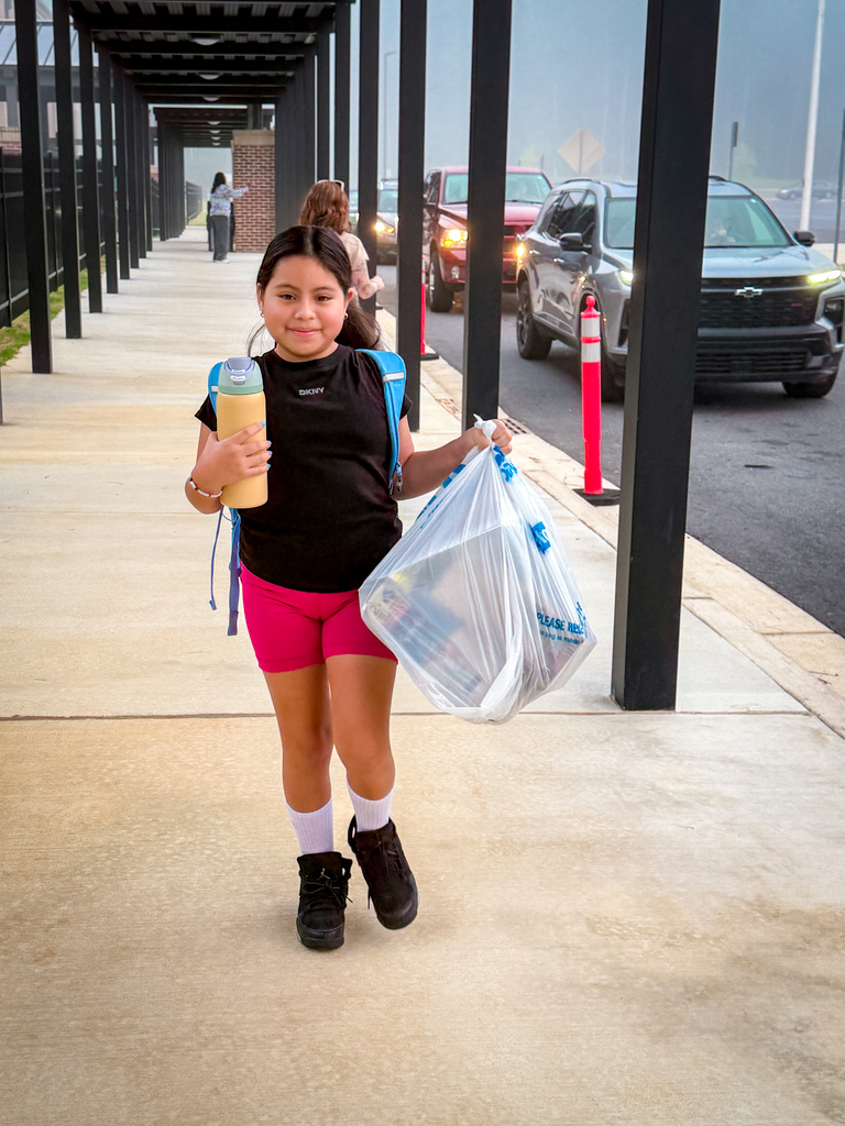 A young girl wearing a black shirt and pink shorts walks on a sidewalk with plastic bags, a car behind.