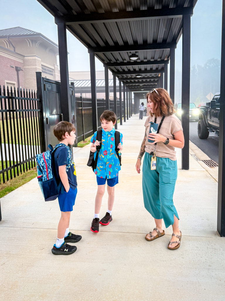 Two children in school uniforms and backpacks stand outside, one looking at a woman holding a phone. The woman stands under a covered walkway.
