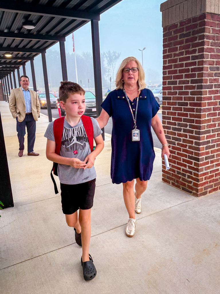 Woman and child walking outside school. Woman holds paper and ID. Child wears backpack. Adult man walks behind.