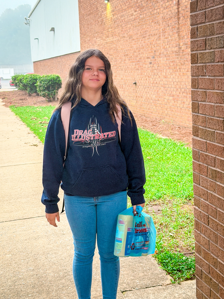 A person in a hoodie and jeans stands on a sidewalk, holding a plastic bag. A brick wall, bushes, and grass are behind them.