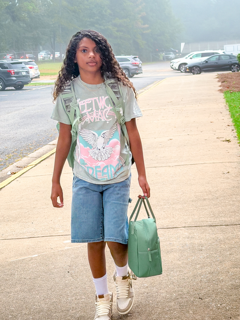 Person wearing denim shorts, white socks, and sneakers walks on a sidewalk, carrying a green bag.