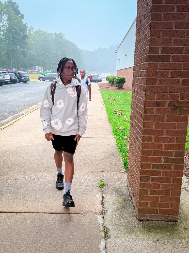 Person in white hoodie, black shorts, and sneakers walking on sidewalk with brick pillar and green grass.