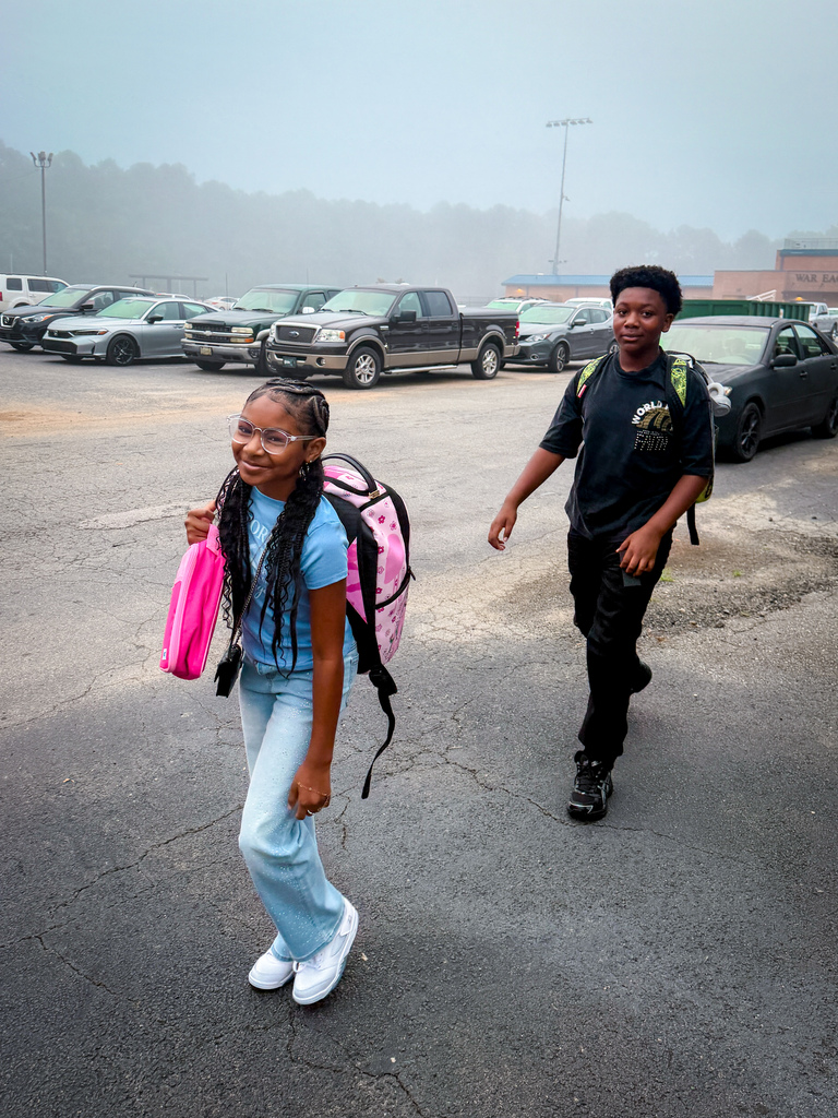 Two children walking through a parking lot with backpacks. They are surrounded by parked cars.