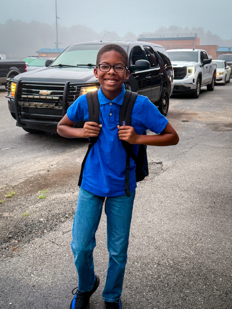 A young person wearing a backpack stands in a parking lot with several vehicles behind them.