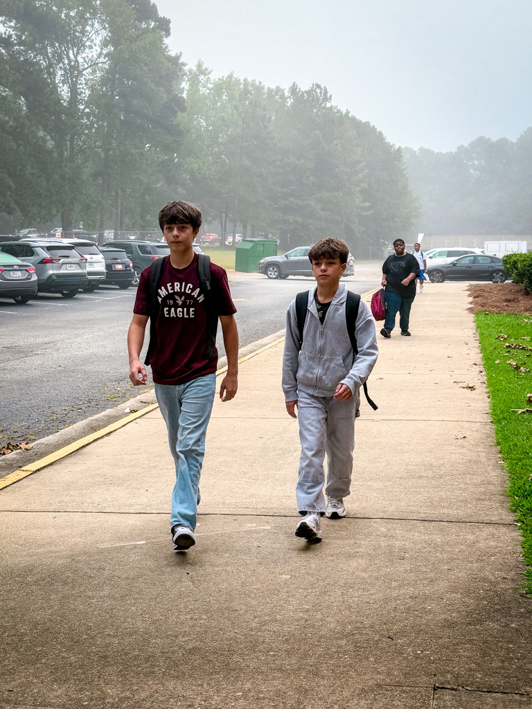 Two boys walk on a sidewalk; one wears a maroon shirt, jeans, and sneakers, the other a gray sweatshirt, pants, and sneakers.