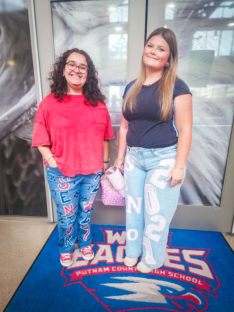 Two women in front of a glass door with a school logo and mascot. One wears red, glasses, and sneakers; the other has long blonde hair and a blue shirt.