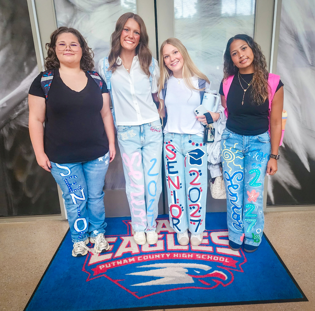 Four women stand on a blue carpet with Putnam County High School Eagles logo. They wear jeans with designs including numbers and years.