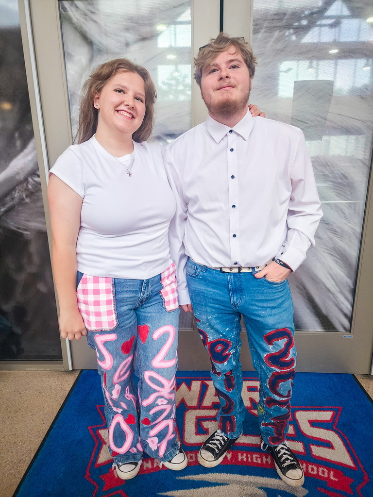 Two people pose for a photo on a rug in front of glass doors. Both wear jeans with art on them.