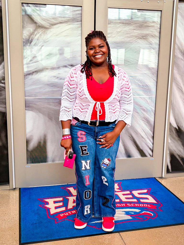 A person in a red top, white cardigan, and blue jeans stands in front of a glass door.