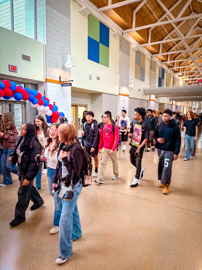 Students wearing various clothing and footwear walk through a brightly lit hallway with colorful balloons.