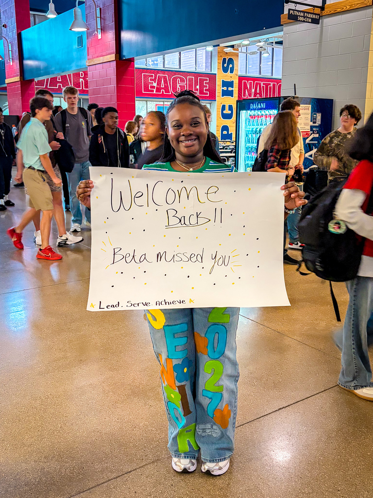 Person holding a white sign saying "Welcome Back!!! Bela missed you" stands in a hallway with other people.
