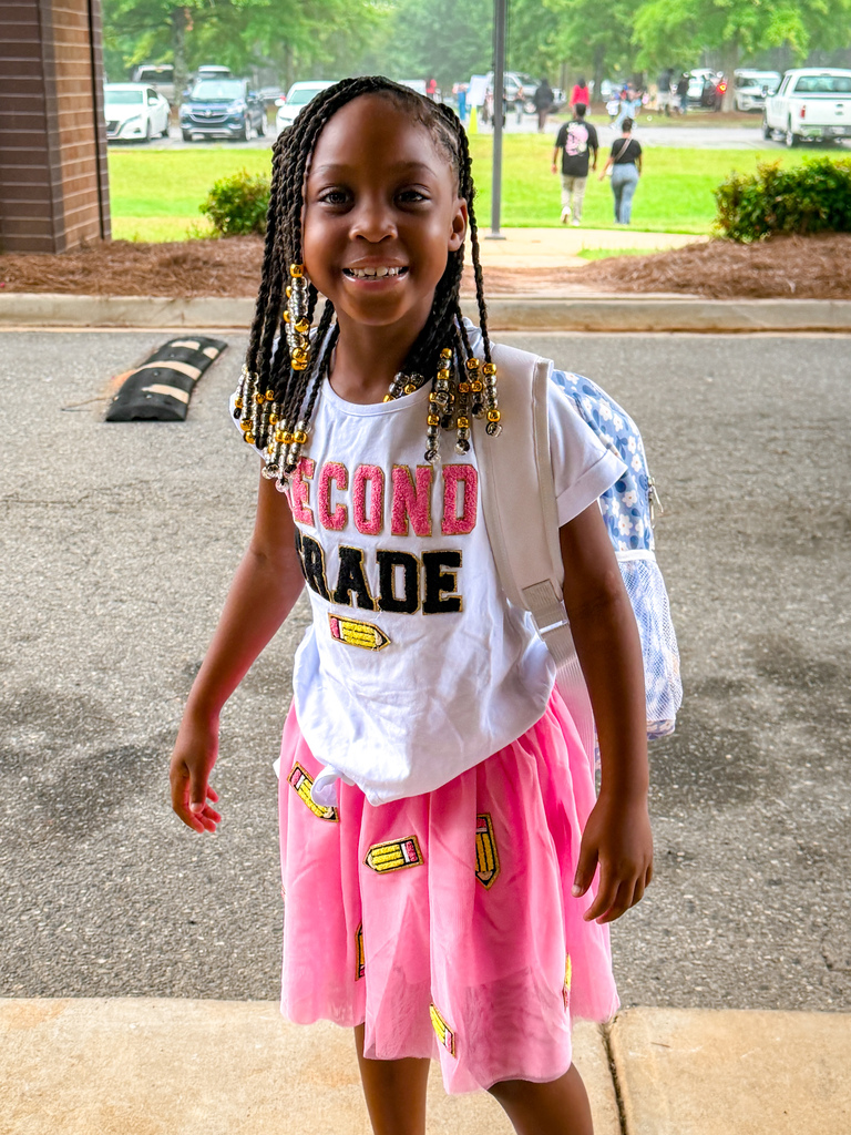 A young girl with braided hair stands in a parking lot. She wears a pink skirt and white top with "SECOND GRADE" in red.