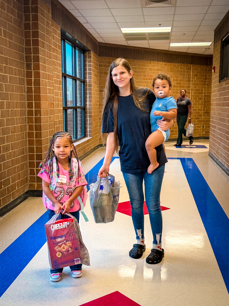 A woman carrying a baby walks in a hallway with two children, one holding a bag.