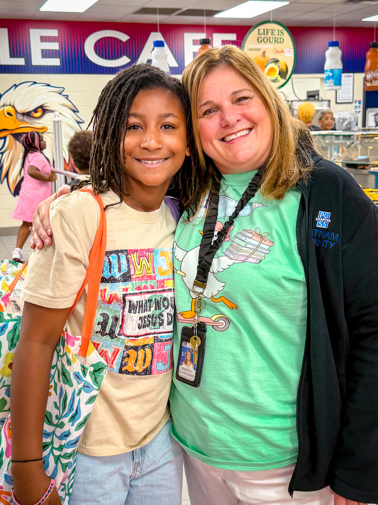 A smiling woman and a young girl pose in a cafeteria, with an eagle and menu boards in the background.