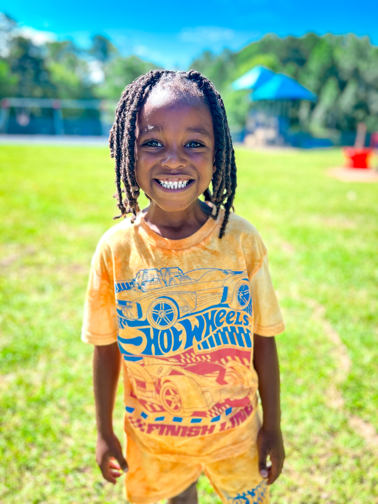 Child with braided hair stands in grassy field, smiling, wearing yellow T-shirt with car design and yellow shorts.