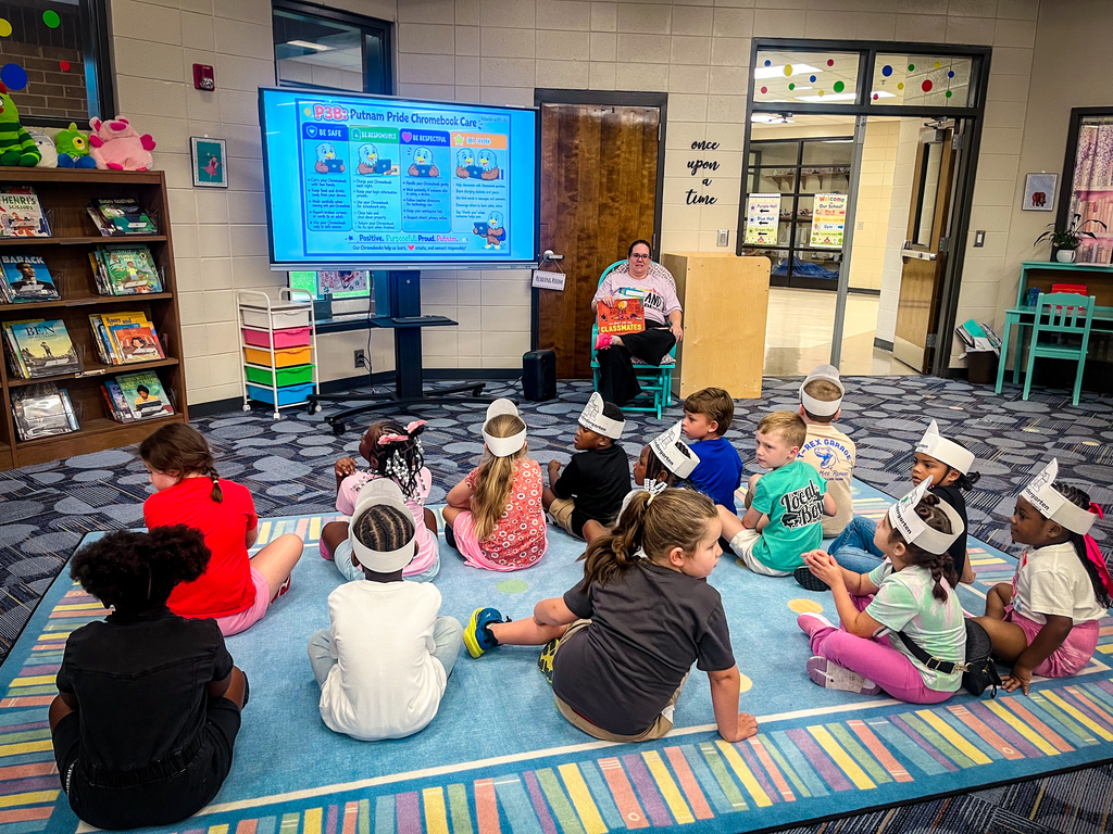 A group of children sit on a rug, facing a screen displaying information. One adult sits in a chair beside them.